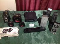 Overview of lot showing DVD player, multiple speakers in various sizes, and boxed Pioneer speaker set on a green tablecloth with maroon curtain background.