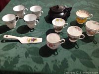 Photo showing a dark brown teapot, nine various floral china tea cups, and a ceramic pie server with purple and yellow flowers on green tablecloth.