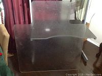 Top view of transparent plastic floor protector mat with visible surface scratches and marks, placed on carpeted floor next to furniture.