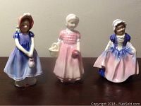Front view of three Royal Doulton girl figurines standing on a surface, showing colors and details of dresses and accessories.