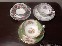 Photo showing all three teacups on their matching saucers with floral designs and gold/silver trims on a wood surface.