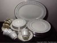 Photo showing large platter, stacks of various plates including dinner, salad, and bread plates, plus teacups and saucers arranged on a flat surface in soft lighting