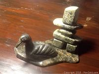 View of signed Inuit soapstone carving showing bird figure on base with stacked geometric stone man figure behind it.