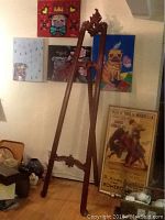 Full view of the tall wooden tripod easel set against a wall with artwork and framed posters around it.