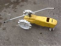 Full side view of the yellow cast iron tractor-shaped water sprinkler showing large white spiked wheels, metal handle, and watering outlet on top.