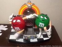 Front angle showing red and green M&M characters with arms raised beside an orange-yellow accented jukebox on a black and white checkered base.