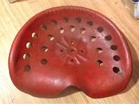 Top view of a red-painted metal tractor seat with ventilation holes and "MASSEY HARRIS" embossed.