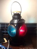 Front view showing the lantern with blue and red glass lenses, with electric cord visible. The red lens is intact, one blue lens is cracked.