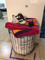Laundry basket filled with towels and topped with iron, hair curler and vanity mirror
