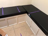 Overall view of corner bench with three heavy duty black cushions attached with purple straps. Bench is made of white painted wood with paneled sides.