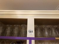 Shelves inside a wooden cabinet holding a row of clear vintage drinking glasses of two sizes with textured rims, top view