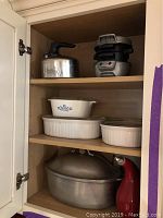 Cabinet shelf with large metal kettle on top shelf, an electric sandwich or panini press, and three ceramic CorningWare dishes on second shelf; a large oval metal Dutch oven with lid on bottom shelf.