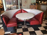Front angle showing the red and white vinyl upholstery of the corner booth with black trim, along with the round white tabletop and chrome base.