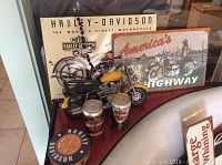 Photo showing Harley Davidson replica motorcycle, two metal signs, collectible cans, and coaster tin on display