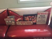 Five novelty metal signs displayed on a red booth seat including diner and poker themes, plus one framed electric art picture leaning on the booth.