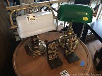 Two banker style desk lamps and two wooden rock-decorated bookends on round wooden table.