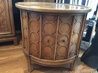 Front view of round wooden side table with carved circular motifs on cabinet doors and wooden handles.