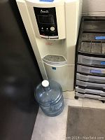 Full height photo of off-white Avanti water dispenser with a large empty water bottle on the floor beside it.