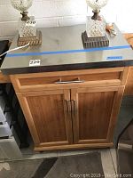 Kitchen cart with wooden frame and metal top. Two glass lamps on the top surface. Drawer and double door cabinet visible.
