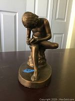 Front view of 9-inch nude statue seated with one leg crossed, hands examining foot.