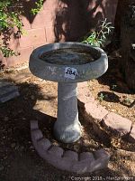Concrete bird bath set up outdoors on soil near a tree and brick wall, showing full view, two-piece construction, and surrounding stone border.