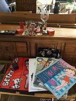 Wide photo showing an assortment of Betty Boop collectibles on a dresser including tins, shot glasses, posters, boxes and wallet