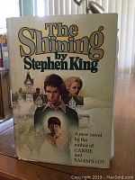 Front cover of hardcover book The Shining by Stephen King showing illustrated dust jacket with faces, dog, and building.