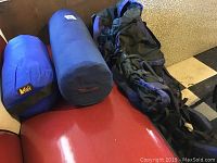 Photo of two blue stuff sacks containing a sleeping bag and a camping mattress, and a dark blue and black framed backpack on a red bench, showing surface dust and use.