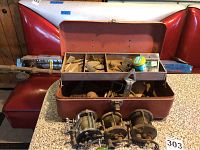 Photo showing vintage metal tackle box open with assorted fishing lures inside and three fishing reels in front.
