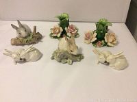 Full view of all porcelain items together: two Capodimonte candleholders with roses and greenery, two Lladro white bunny figurines, and two smaller Denmark bunny figurines sharing lettuce.