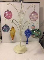 Overall view of 8 hand blown glass ornaments hanging on cream metal display stand.