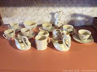 Overall assortment arranged on countertop showing five teacup/saucer sets, two swan vessels, creamer, square dish, and tall ewer