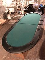 Green felt oval poker table with black padded edges and multiple cup holders, placed on cardboard boxes in a basement-like storage area.