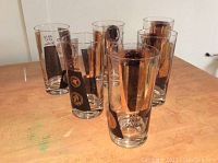 Photo showing six vintage highball glasses arranged on a wooden surface, each decorated with a currency motif featuring coins and dates in gold and black print.