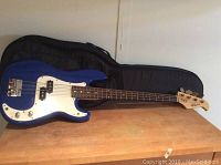 Full side view of the blue Kima electric bass guitar laid on a wooden surface with the black Pro Tec carrying case open behind it.