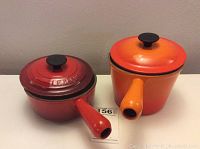 Pair of Le Creuset sauce pans side by side on a flat surface showing orange and red exterior colors, with lids on top, black knob handles, hollow handles visible, made in France.