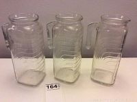 Set of three tall clear glass juice jars with ribbed patterns and handles, shown side by side