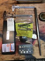 Various jeweler's tools laid out on a wooden surface including calipers, ring gauge, compass and small parts in bag.