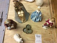 Six ceramic figurines and ashtray shown on wooden surface, including religious, Hummel children, and Doulton style older woman.