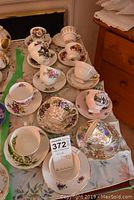 Top view showing twelve teacups and saucers arranged on table, displaying assorted floral patterns and styles