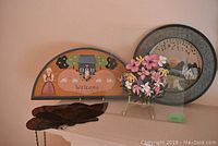 Photo of folk art painted wood half-moon plate with house and floral welcome design, round floral painted metal plate, and pink artificial flowers on display.