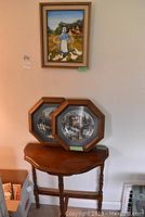 Wood demi lune table with two framed plates on top and a framed painting hanging above on the wall, showing arrangement of the lot