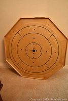 Full view of an octagonal wooden crokinole board placed against wall on carpeted floor, showing scoring rings and numbers.