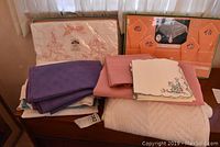 Photo of stacked vintage linens showing boxed 1970s tablecloth and napkin sets with embroidered floral designs, various purple and pink folded textiles, and white quilted bedding.