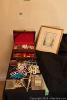 Photo showing open wooden Princess Royal jewellery box with red interior housing various costume jewellery pieces including bracelets, watches, brooches and necklaces, and a framed print propped nearby.