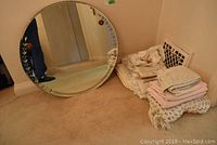 Photo shows round glass mirror with leaf and rose decorative border placed against a wall near a pile of folded linens including bath mat, afghan, and fabric items.
