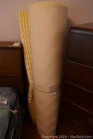 Foam mattress topper rolled up and placed upright next to furniture, showing full height and flexibility.