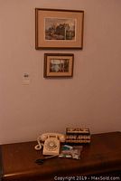 Wide shot showing rotary dial phone, sewing box, and two framed prints on wall above a wooden surface
