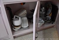 Photo showing cupboard with white electric hand mixer, plastic Tupperware, glass baking dishes, metal pots, and baking trays