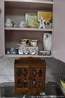 Photo showing kitchen cabinet interior with assorted small kitchen helpers: salt and pepper shakers, cream and sugar sets, cookie and noodle maker, can opener, above hardwood spice rack.